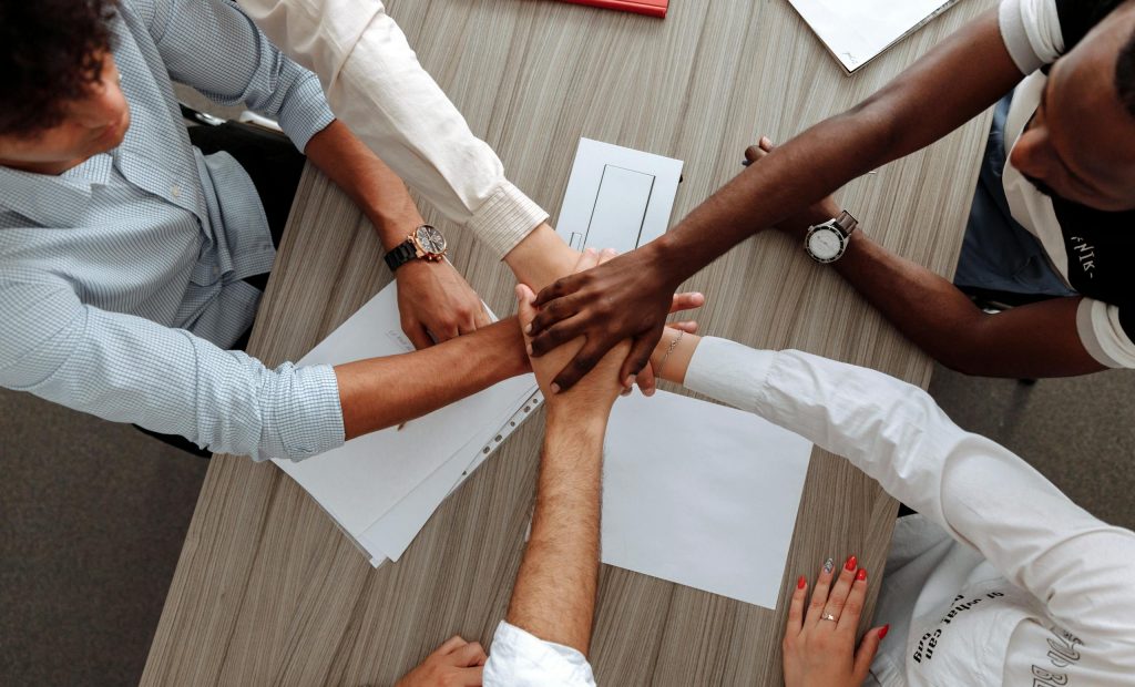 A diverse group of adults join hands in an office, symbolizing teamwork and cooperation.